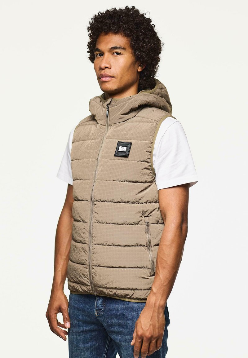 Beige gepolsterte Weste mit Kapuze, mit einem Reißverschluss vorne, Seitentaschen und einem kleinen schwarzen Logo-Patch. Getragen über einem weißen Hemd.
