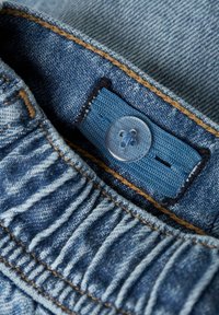 Vêtement en denim avec un bouton bleu et une étiquette en tissu présentant un motif brodé. La taille a une texture froncée et des coutures beiges.