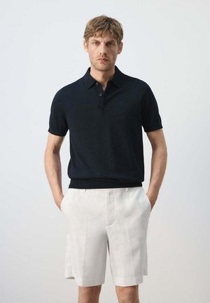 Hombre de pie con las manos en los bolsillos, lleva una camisa polo negra de manga corta y pantalones cortos blancos hasta la rodilla sobre un fondo liso.