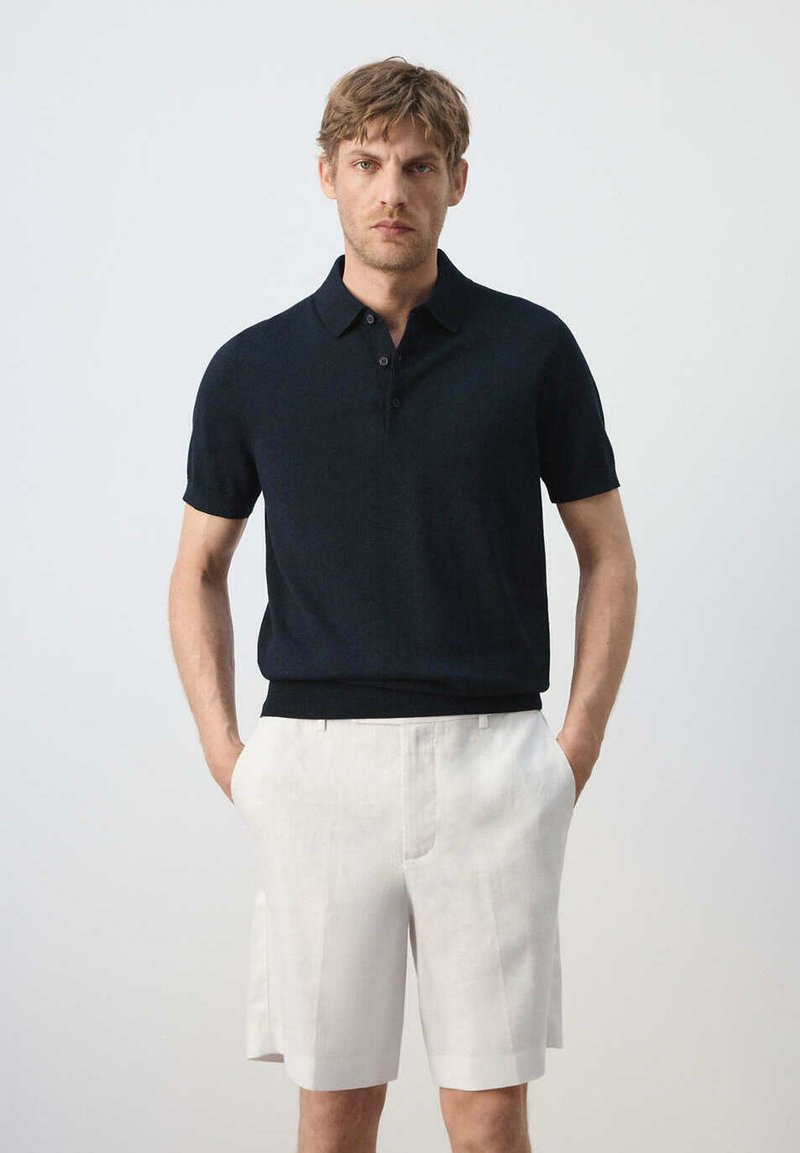 Homme debout les mains dans les poches portant un polo noir à manches courtes et un short blanc jusqu'aux genoux sur fond uni.