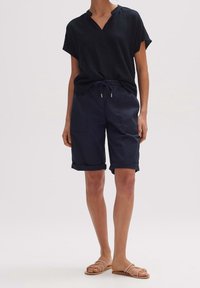 Granatowa bluzka z krótkim rękawem o teksturowanym materiale z dekoltem w serek, w zestawie z granatowymi szortami z podwiniętymi nogawkami, posiadającymi kieszenie i sznurek, noszona z beżowymi sandałami.