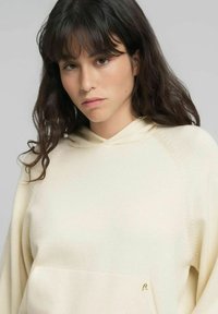 Sudadera con capucha color crema con un bolsillo delantero, presenta una textura sutil y un pequeño logo dorado en la parte inferior izquierda.