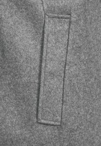 Tejido gris de felpa con una costura visible y un bolsillo de parche rectangular. La textura es suave y ligeramente peluda, enfatizando la calidez.