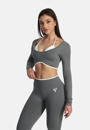 Kvinde i grå langærmet crop top og matchende leggings poserer med den ene hånd på hoften og iført en hvid bikinitop under.