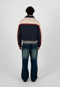 Un homme aux cheveux bouclés et foncés se tient de dos, portant une veste bleu marine, beige et rouge, associée à un jean bleu délavé à jambes larges.