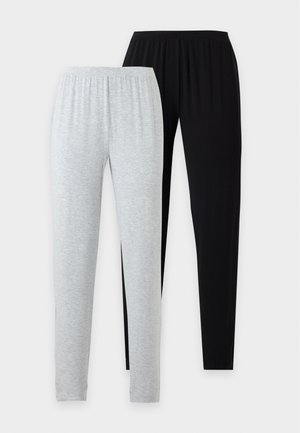 Femme debout pieds nus, portant un haut blanc sans manches et un pantalon noir ample sur un fond clair uni.