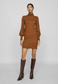 Abito a maglia marrone con collo alto, maniche lunghe a sbuffo, silhouette aderente e texture liscia, abbinato a stivali marroni con tacco al ginocchio.