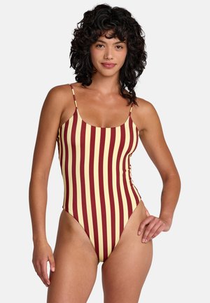 Woman with curly dark hair wearing a one-piece swimsuit with vertical yellow and maroon stripes, standing with one hand on her hip.