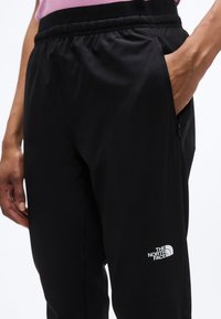 Άτομο που φοράει μαύρο παντελόνι The North Face με ελαστική μέση και το χέρι σε φερμουάρ πλαϊνής τσέπης, φορόντας ροζ μπλούζα.