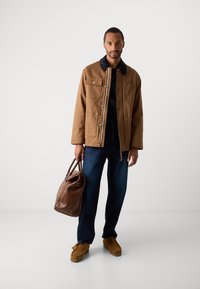 Hombre con chaqueta marrón, jeans oscuros y zapatos marrones a juego sostiene un bolso de cuero marrón mientras está de pie contra un fondo blanco liso.
