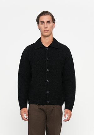 Young man wearing a black buttoned wool cardigan and brown trousers, standing against a plain white background.