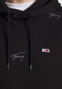 Sudadera negra hecha de tela suave, con estampados de logo en gris y un pequeño logo bordado en rojo, blanco y azul en el frente.