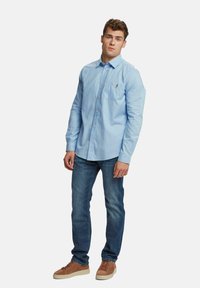 Hellblaues langärmliges Hemd mit Knopfleiste und Brusttasche, kombiniert mit dunkelblauen Jeans und braunen Wildledersneakern. Einfaches, lässiges Design.