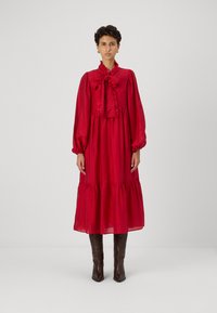 Piros, hosszú ujjú ruha fodros gallérral és megkötő részlettel, rétegzett szoknya és széles ujjak. Térd felettig érő barna csizmával viselve.