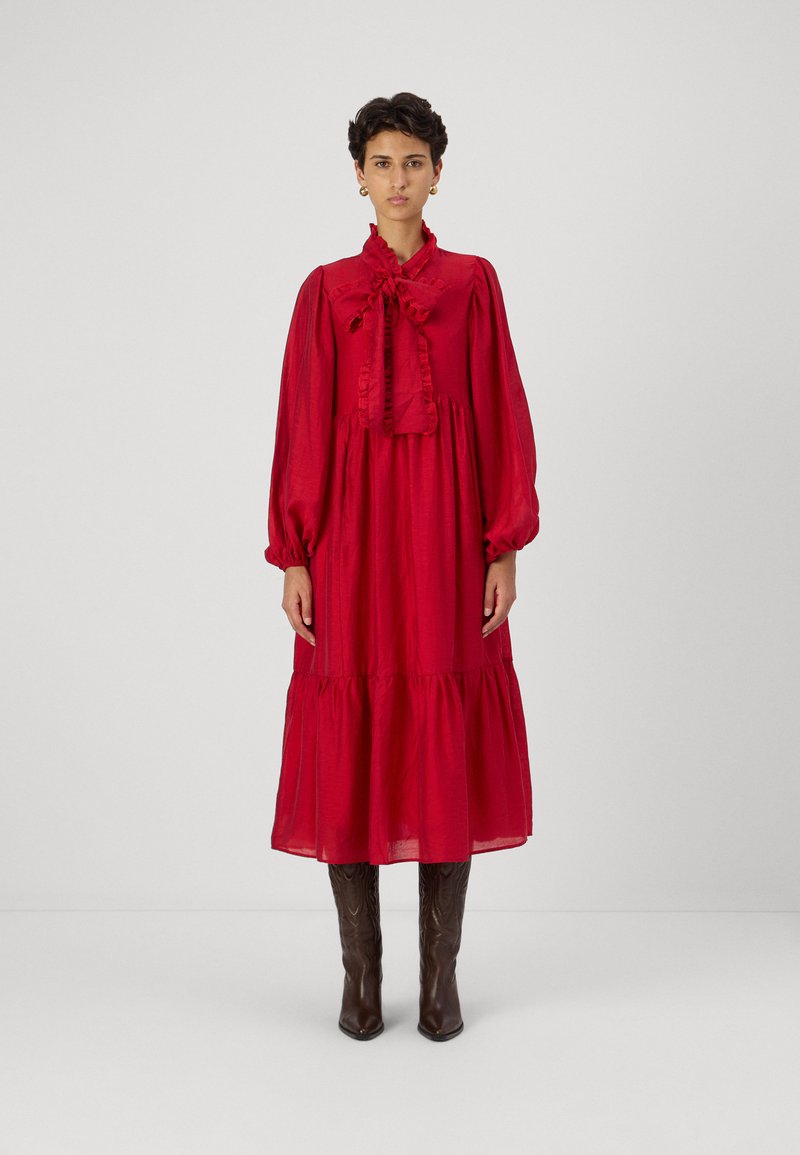 Piros, hosszú ujjú ruha fodros gallérral és megkötő részlettel, rétegzett szoknya és széles ujjak. Térd felettig érő barna csizmával viselve.