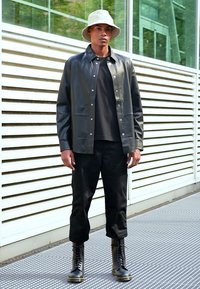 Camisa chaqueta de cuero negro con botones a presión, llevada sobre una camiseta negra, combinada con pantalones negros y botas de cordones negras. Sombrero bucket blanco.