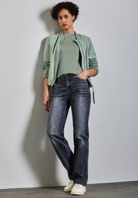 Světlá zelená bomber bunda přes šedé tričko, v kombinaci s tmavými džínami a bílými teniskami. Hladká látka, volný střih a jemné textury.