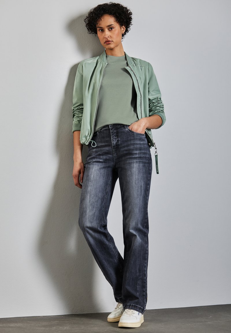 Světlá zelená bomber bunda přes šedé tričko, v kombinaci s tmavými džínami a bílými teniskami. Hladká látka, volný střih a jemné textury.