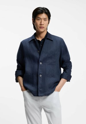 Jonge man draagt een donkerblauwe jas met knopen en opgerolde mouwen en witte broek, staand met handen in de zakken tegen een witte achtergrond.