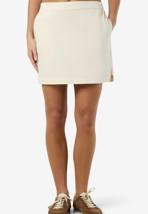 Kobieta w beżowej spódnicy do kolan z bocznymi kieszeniami i brązowych zamszowych trampkach z białymi sznurowadłami, stojąca na tle białego tła.