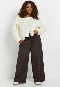 Ivory gebreide cardigan met grote knopen, gecombineerd met brede bruine broeken. Beide stukken hebben een ontspannen pasvorm en opmerkelijke zakken.
