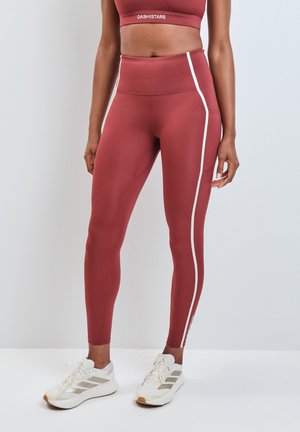 Donna che indossa leggings alto vita di colore marrone rossiccio con strisce laterali bianche e scarpe da ginnastica bianche, in piedi su uno sfondo chiaro e uniforme.