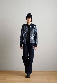 Veste bomber noire avec des motifs abstraits bleus, associée à un jean foncé orné de broderies rouges. Le modèle porte un bonnet noir.