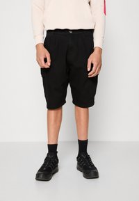 Czarne shorts cargo z bawełny z bocznymi kieszeniami, luźny krój i podwinięte nogawki, w połączeniu z czarnymi butami sportowymi i czarnymi skarpetkami.