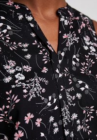 Schwarze Bluse mit floralem Muster, die eine Mischung aus rosa und weißen Blumen zeigt, kragenlosem Ausschnitt und einem leichten Stoff mit glatter Textur.