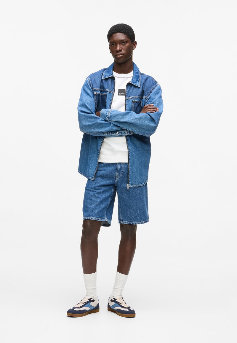 Jeansjacke mit blauem Waschung, kurze Jeansshorts, weißes T-Shirt mit Logo und mehrfarbige Turnschuhe. Weiße Crew-Socken sichtbar.