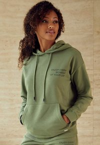 Olivgrüne Kapuzenjacke aus weichem Stoff, mit einer Vordertasche im Känguru-Stil, einer Kordelzugkapuze und bedrucktem Text: „GENIEßE JEDEN MOMENT DEINES LEBENS.“