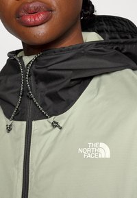 Jasnozielona kurtka z czarnym kapturem, z przodu zapinana na zamek błyskawiczny i z regulowanym sznurkiem. Posiada białe logo po lewej stronie klatki piersiowej. Wykonana z gładkiego materiału.