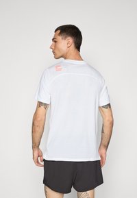 Hvid atletisk t-shirt med korte ærmer, der har en mesh-struktur og et rødt trykt logo på den øvre ryg. Parret med sorte shorts.