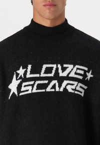 Μαύρο πλεκτό πουλόβερ με ψηλό γιακά, που φέρει λευκή επιγραφή "LOVE SCARS" μαζί με αστέρια στη διακόσμηση.