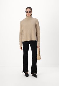 Pull turtleneck côtelé beige à manches longues, associé à un pantalon noir évasé. Le modèle tient un petit sac à main beige et porte des lunettes de soleil foncées.