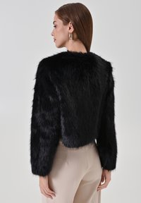 Veste courte en fausse fourrure noire à manches longues. Caractérisée par une texture douce et des épaules arrondies, associée à un pantalon ajusté de couleur claire.