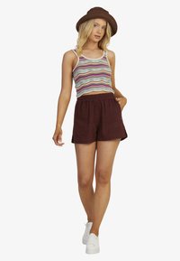 Stribet afkortet tanktop i blå, elfenben og maroon; parret med høj talje bordeaux shorts; modellen bærer hvide sneakers og en brun hat.