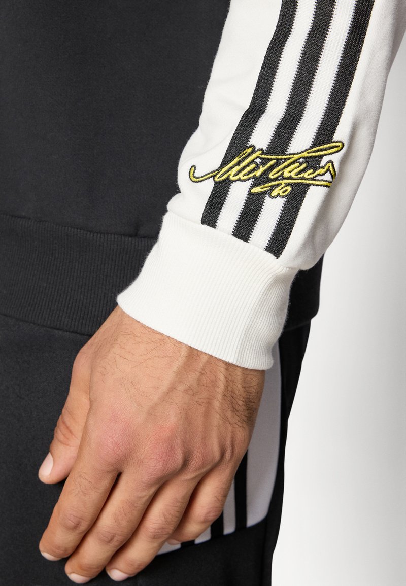 Schwarzer und weißer Sweatshirtärmel mit drei vertikalen Streifen und gesticktem gelbem Signaturdetail am gerippten Bündchen.