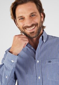 Camisa de botones azul y blanca a rayas con cuello, que cuenta con un bolsillo y un logo en el lado izquierdo del pecho, confeccionada en tejido ligero.