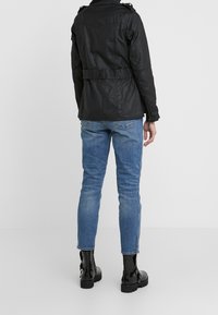 Schwarze wasserdichte Jacke mit tailliertem Gürtel, Schulterklappen und geradem Design, kombiniert mit blauen Jeans und schwarzen Knöchelstiefeln.