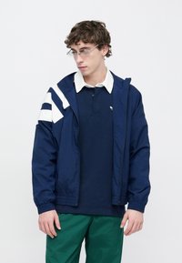 Tengerészkék dzseki fehér cikk-cakk mintával, elöl cipzárral és elasztikus mandzsettával; alatta kék galléros póló és zöld nadrág. Átlátszó szemüveget visel.