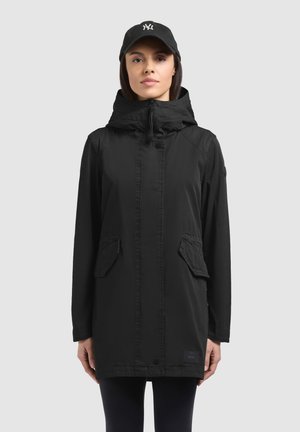 Nő fekete kapucnis dzsekiben, zsebekkel, és fekete baseball sapkában fehér logóval, szürke háttér előtt áll.