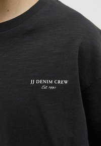 Melnas kreklu arapa veidotā T-kreklu, izgatavots no mīkstas auduma, ar baltu uzrakstu "JJ DENIM CREW" un "Est. 1990" kreisajā krūtīs.