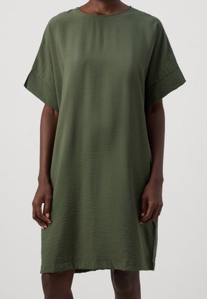 Vestito verde oliva a maniche corte, largo, con ampi polsini, indossato da una persona in piedi davanti a uno sfondo chiaro e uniforme.