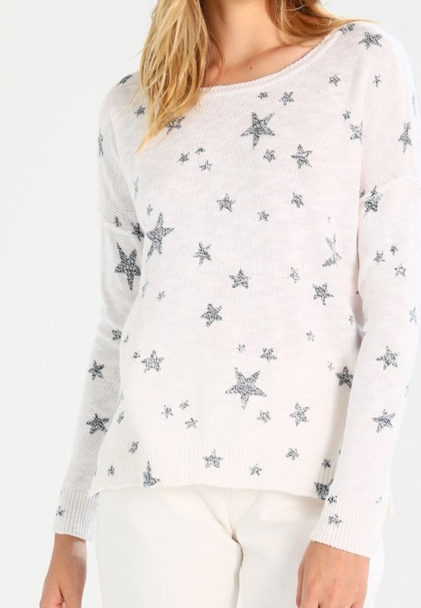 Kvinna iklädd en vit stickad tröja med spridda grå stjärnmönster, i kombination med vita byxor, visad från axlarna till höfterna.