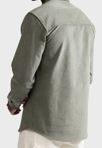 Homme portant une chemise en velours côtelé vert olive clair à manches longues vu de dos avec un pantalon beige sur fond uni.