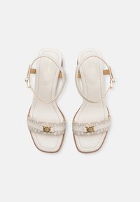 Beige hælede sandaler med en struktureret rem, der har en guld logo detalje. Remme omslutter anklen med et firkantet tådesign.