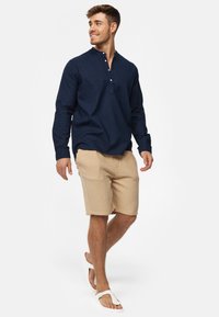 Marinblå långärmad skjorta med knappad halsringning, tillsammans med beige texturerade shorts och vita flip-flop-sandaler, på en modell som går.
