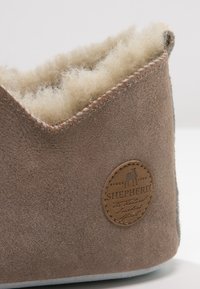Brown suede slipper with a soft cream shearling lining, low-cut design, and a circular tan leather logo patch. Accented stitching visible.