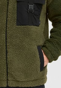 Olive green fleece jacket with a textured surface, featuring a black nylon chest pocket and contrasting black trim along the zipper and cuffs.